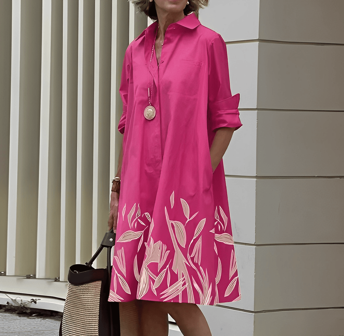 Frau in pinkem Hemdkleid mit floralem Muster, trägt lange Halskette, steht vor moderner Architektur, hält eine Tasche. Elegante Damenmode, Sommerlook.