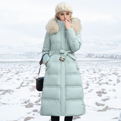 Frau in mintgrünem Daunenmantel mit Fellkapuze steht im verschneiten Winterlandschaft, trägt weiße Mütze und schwarze Tasche. Wintermode, Outdoor.