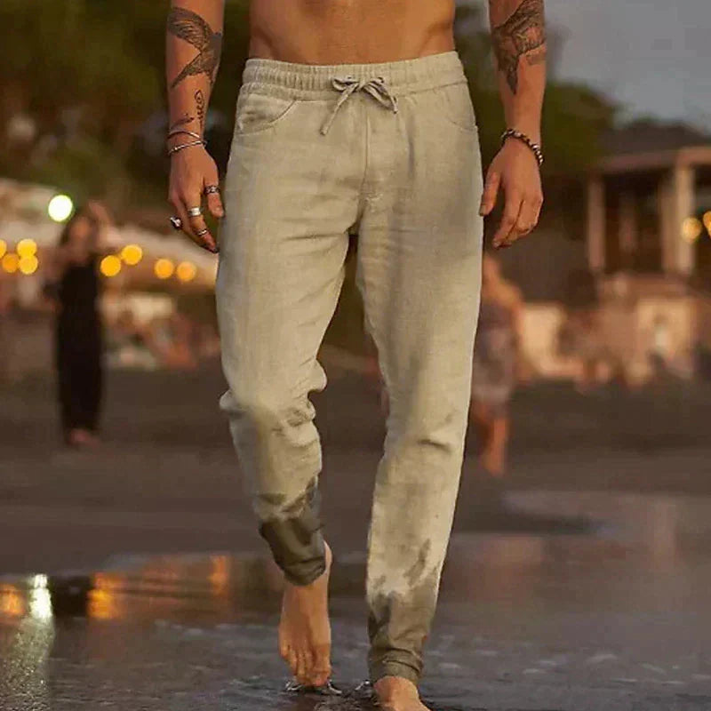 Mann in beigen Jogginghosen am Strand bei Sonnenuntergang, nasse Hosenbeine, lässige Sommermode, barfuß im Sand, entspannte Abendstimmung.