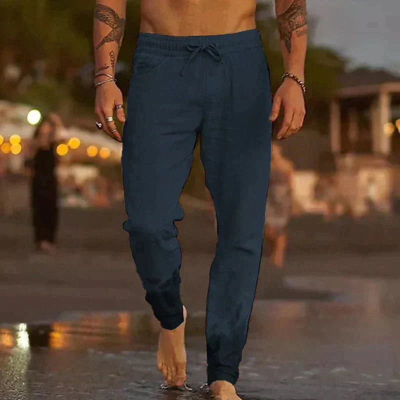 Mann in blauer Jogginghose geht barfuß am Strand entlang bei Sonnenuntergang, Freizeitmode, Sommerabend, entspannte Atmosphäre.