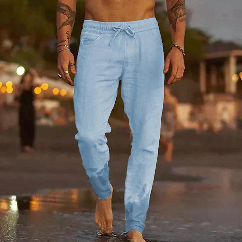 Mann in hellblauer Jogginghose am Strand, barfuß im Wasser, abendliche Strandkulisse. Freizeitmode, Sommerkleidung, Herrenbekleidung.