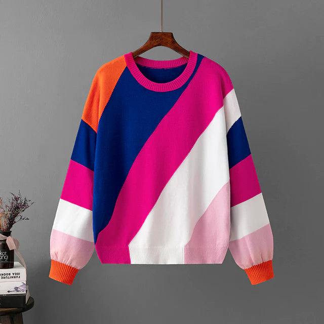 Bunter Pullover mit geometrischem Muster in Blau, Pink, Weiß und Orange, hängend an einem Kleiderbügel vor grauem Hintergrund. Modetrend Herbst.