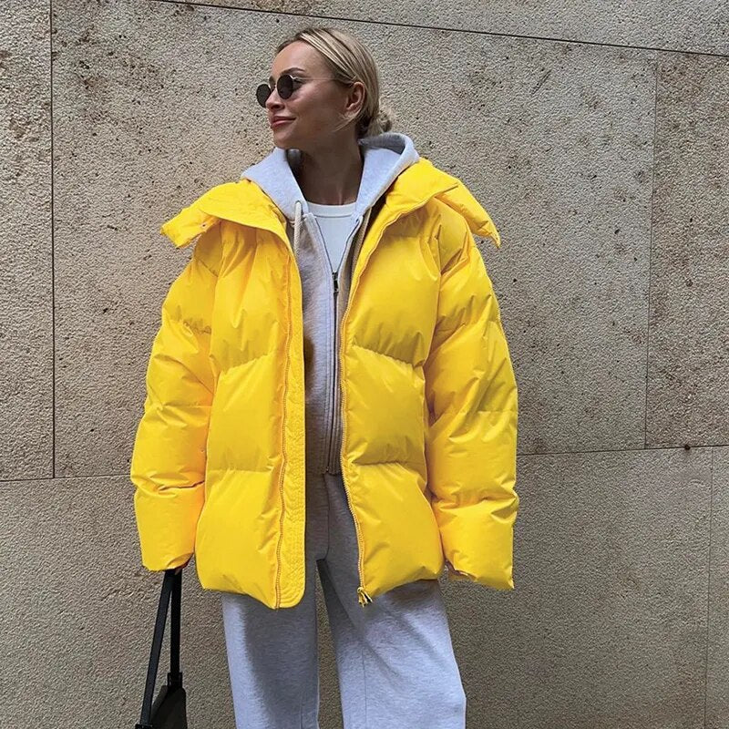 Frau in gelber Daunenjacke, grauem Hoodie und Sonnenbrille vor beiger Wand. Wintermode, Streetstyle, trendige Damenbekleidung.
