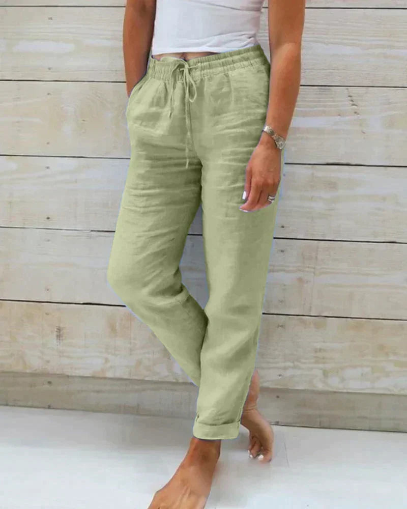 Frau trägt grüne Leinenhose mit Kordelzug, steht barfuß vor Holzwand. Sommermode, lässiger Stil, bequeme Damenbekleidung.