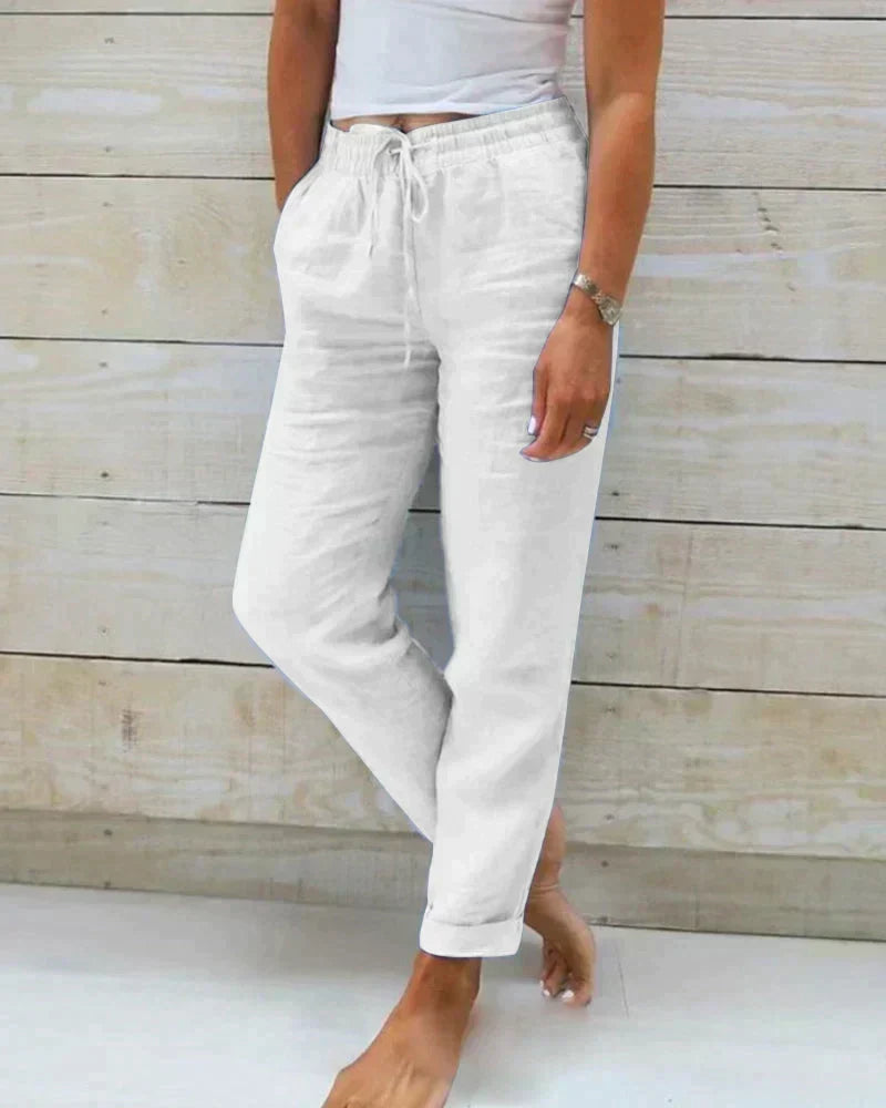 Frau in weißer Leinenhose und weißem Top vor Holzwand, lässiger Sommerlook, bequeme Damenmode, Freizeitkleidung, modische Leinenhosen.