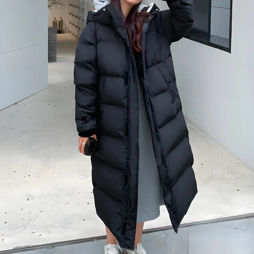 Person in langem, schwarzem Daunenmantel, hält eine Kamera. Wintermode, Outdoor-Bekleidung, stilvoller Mantel, warme Jacke, modischer Look.