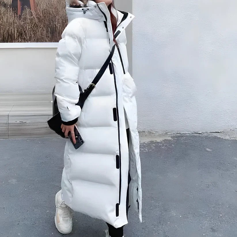 Weiße lange Daunenjacke mit Kapuze, schwarze Akzente, getragen von Person mit schwarzer Umhängetasche und weißen Sneakers, urbaner Hintergrund.