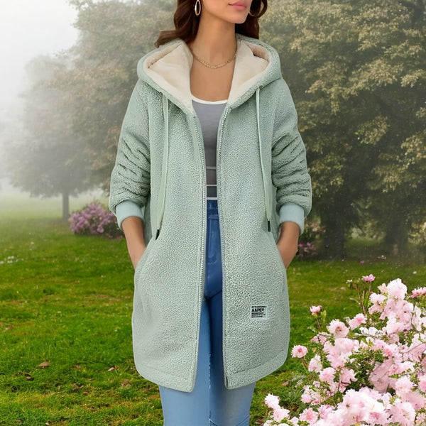 Frau in mintgrünem Teddy-Mantel mit Kapuze, Jeans und weißem Top steht im blühenden Garten. Modetrend, Herbstmode, Damenbekleidung.