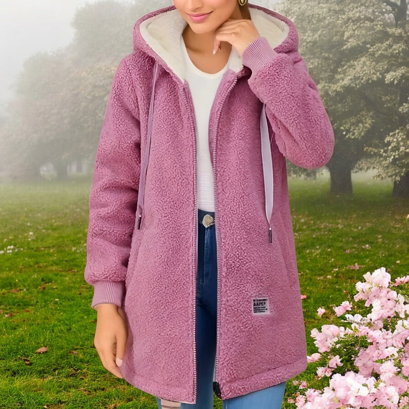 Frau in rosa Teddyfellmantel mit Kapuze, weißem Shirt und blauer Jeans steht im grünen Park. Modetrend Herbst, gemütliche Damenbekleidung.
